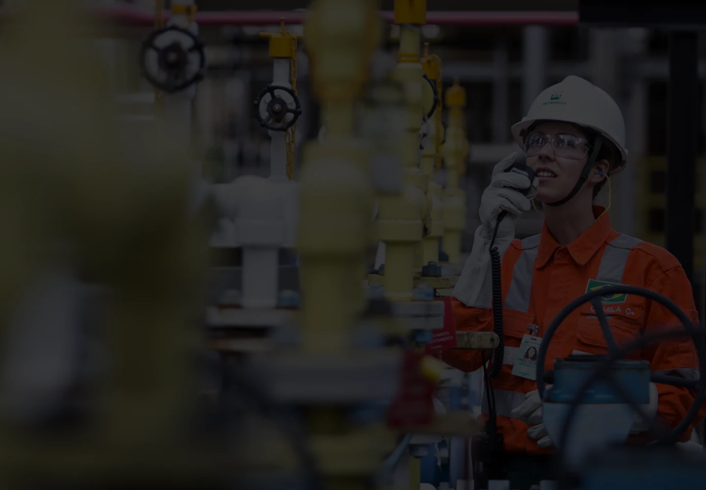 Fotografia diurna de uma funcionária da Petrobras, usando equipamento de proteção completo, falando a um walkie-talkie.