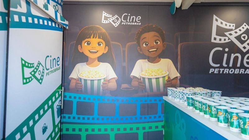 Estande do Cine Petrobras com painel ilustrado de duas crianças sorridentes segurando pipoca, copos temáticos sobre balcão e logomarca da campanha ao fundo. 