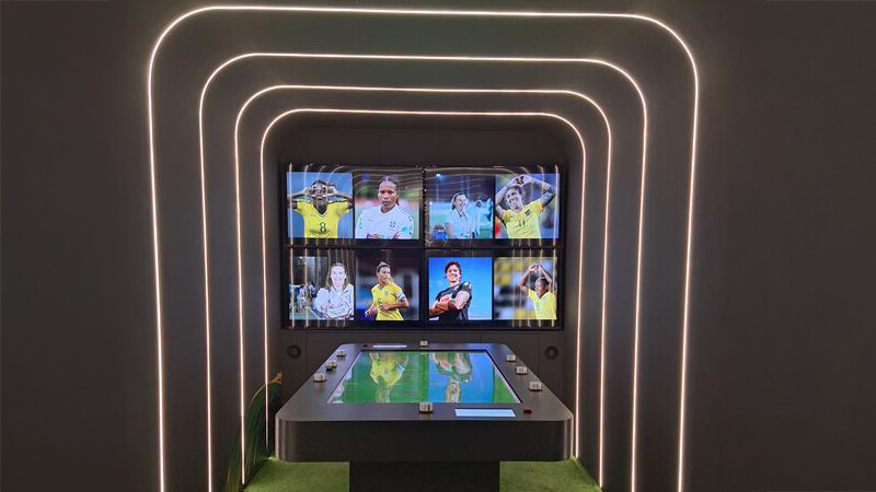 Uma mesa com uma tela no centro e botões ao redor, dentro de uma sala pequena e escura, em frente a um telão com fotos de jogadoras.
