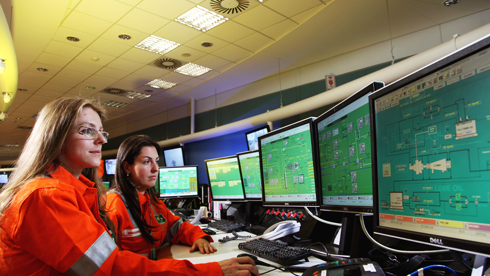 Duas mulheres com uniforme laranja da Petrobras, sentadas em frente a vários monitores.