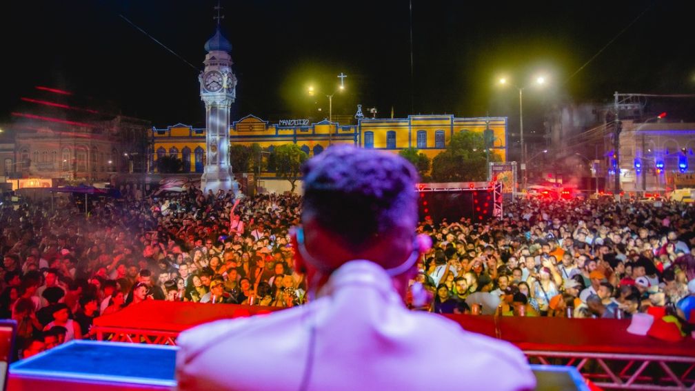 Multidão acompanha show noturno em praça histórica, vista a partir do palco com artista em primeiro plano.