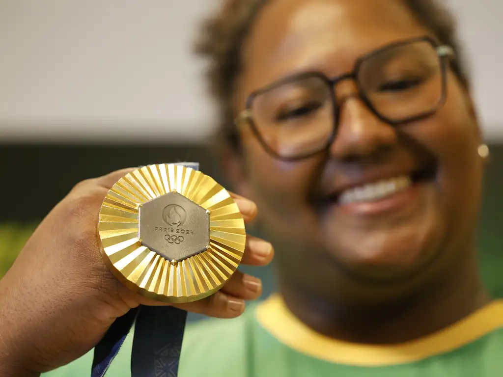 Atleta Bia Souza sorri para a foto segurando uma medalha olímpica de ouro ao lado do rosto.