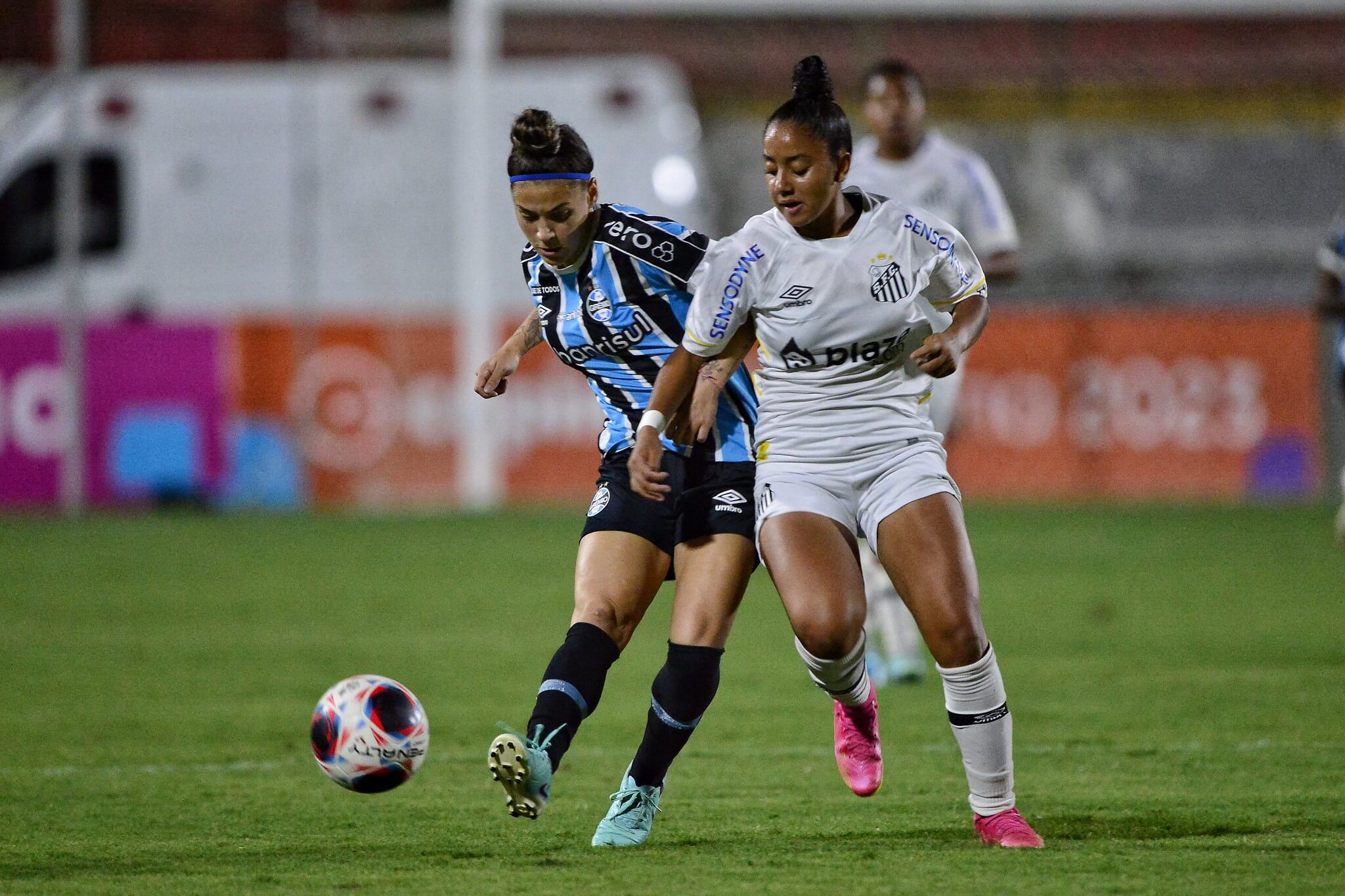 Jogadora do Grêmio chuta a bola, enquanto jogadora do Santos tenta bloquear em jogo da Copinha Feminina de futebol.