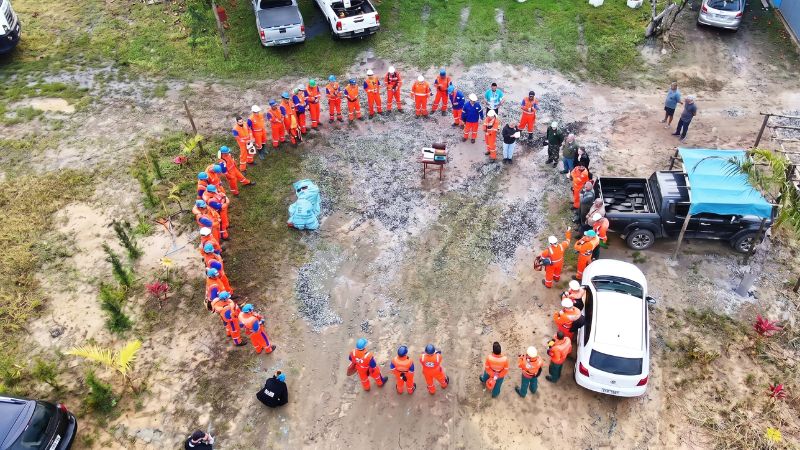 Vista aérea de equipe de campo reunida em semicírculo ao ar livre, com trabalhadores usando macacões laranja e capacetes, em área de trabalho com veículos ao redor. 