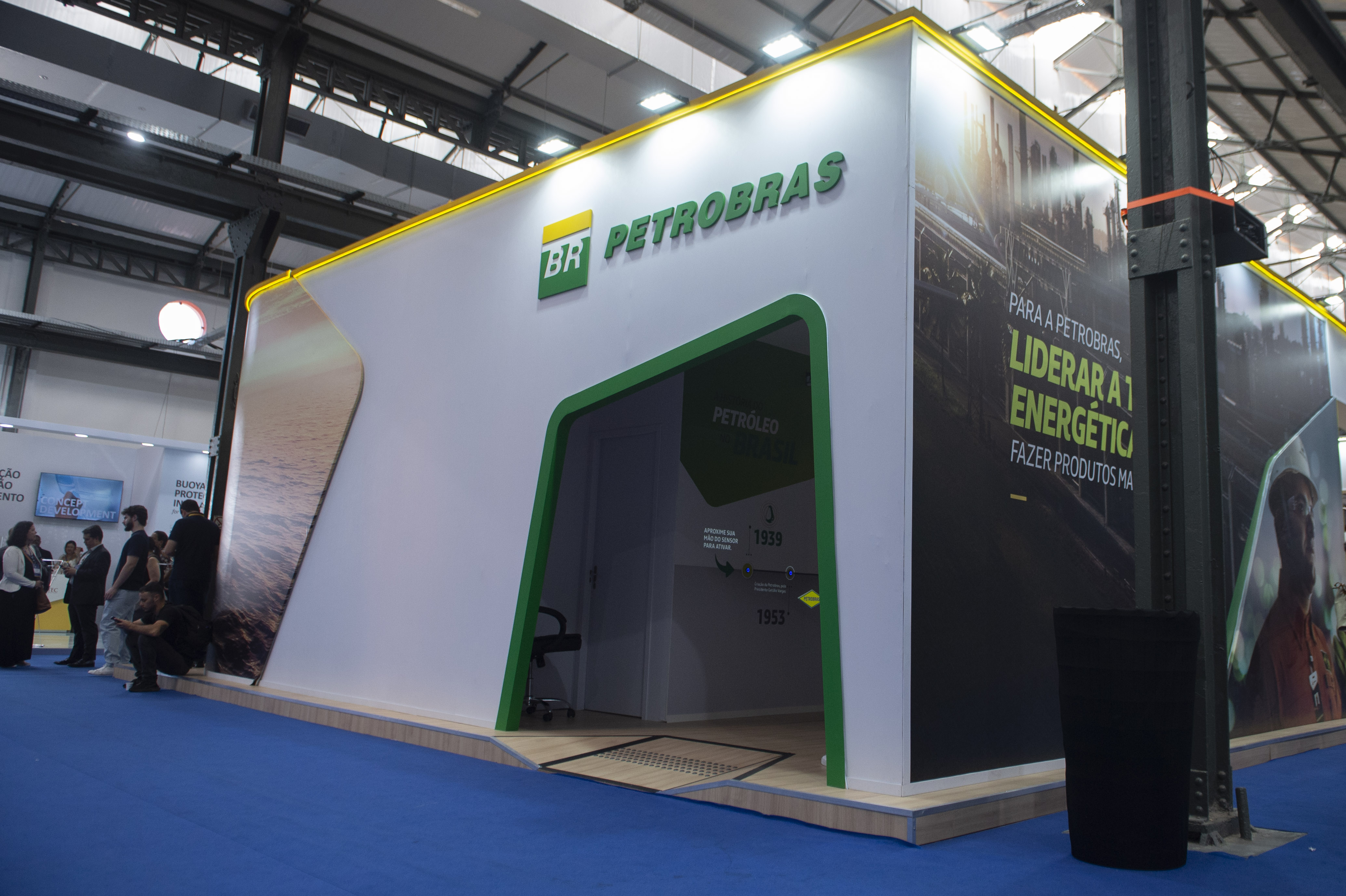 Vista de um estande moderno da Petrobras no evento Rio Oil and Gas, na edição de 2024. A estrutura branca e detalhes em verde e iluminação em LED amarelo no teto. Na fachada principal, destaca-se o logotipo verde e amarelo da marca. O painel lateral exibe um banner preto com dizeres sobre liderar a transição energética. Pessoas caminham e interagem no entorno.