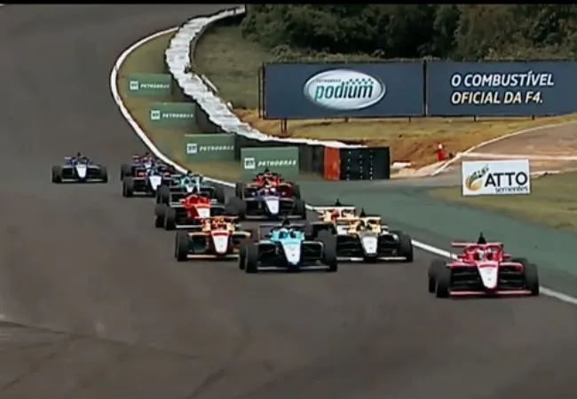 Carros de corrida da Fórmula 4 (F4 Brasil) em uma curva em uma pista, alinhados em fila, com uma placa de publicidade ao fundo indicando Petrobras Podium - O Combustível Oficial da F4.