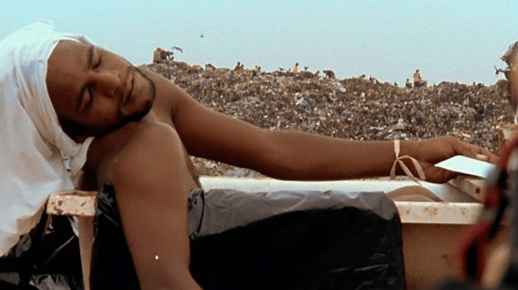 Homem de olhos fechados e com aparência cansada, sem camisa e comum tecico na cama, em meio a um lixão.