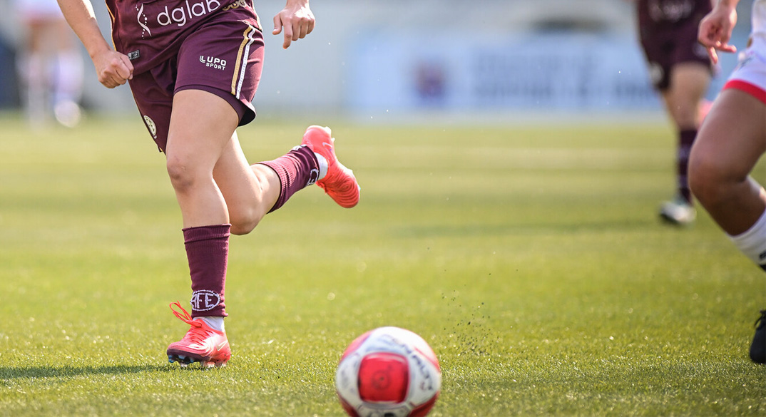 Fotografia diurna mostrando as pernas de duas jogadoras de futebol correndo atrás de uma bola em um gramado.