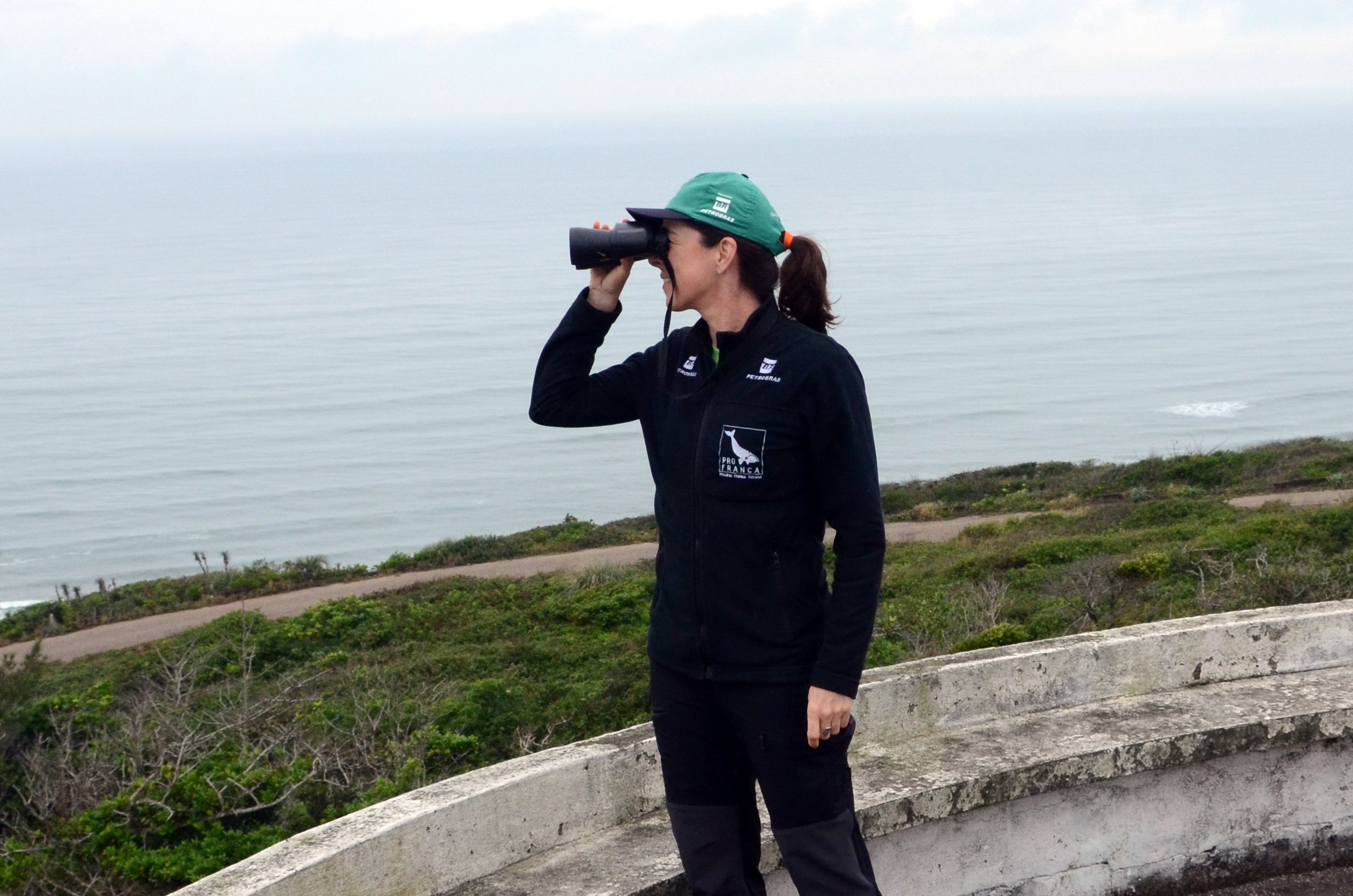 Foto de Karina Groch. Ela é branca, usa boné e está observando o mar com um binóculos.
