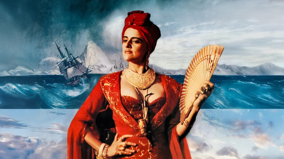 A atriz Marieta Severo, como Carlota Joaquina, em traje vermelho, com um leque na mão, em frente a uma paisagem que divide um mar tempestuoso com um navio e montanhas de gelo na parte superior e um céu com nuvens na parte inferior.