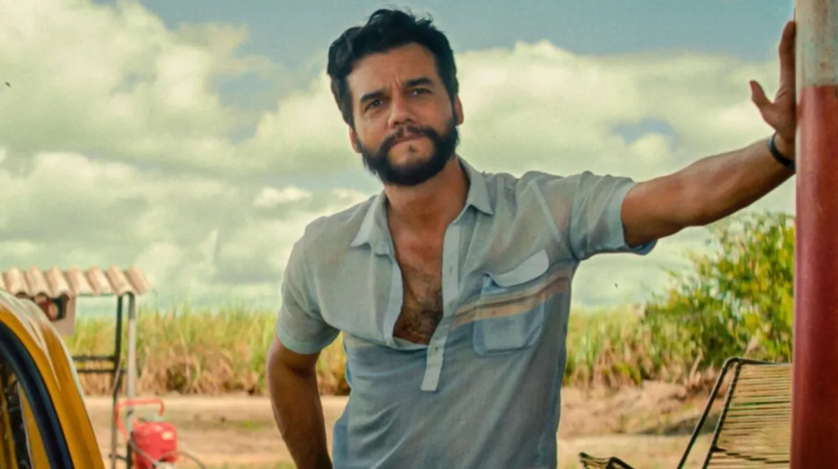 O ator Wagner Moura, usando uma camisa azul clara parcialmente desabotoada, está encostado em um poste vermelho em um cenário rural com um canavial ao fundo e um céu parcialmente nublado.