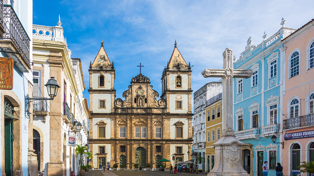 Igreja barroca no Pelourinho, em Salvador, com casas coloridas e cruz de pedra ao centro.