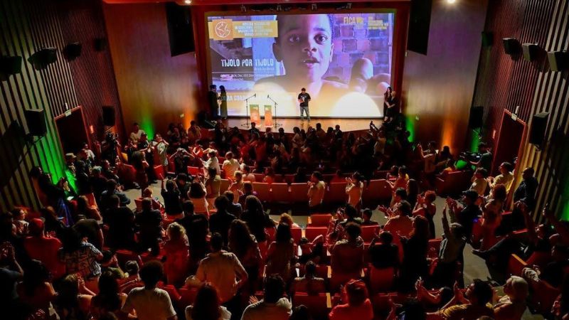 Sala de cinema pequena repleta de pessoas, muitas em pé e batendo palmas.