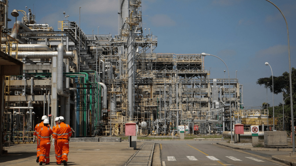 Fotografia diurna do interior da Refinaria Henrique Lage. Quatro homens com uniforme laranja caminham por ela.
