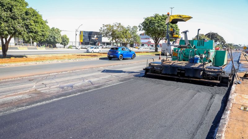 Máquina pavimentadora aplicando asfalto em via urbana, com carros circulando ao fundo.