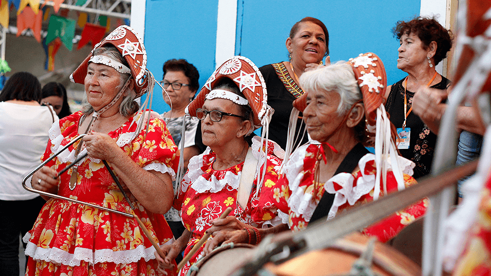 Três mulheres idosas celebram São João com roupas típicas e tocando instrumentos musicais.
