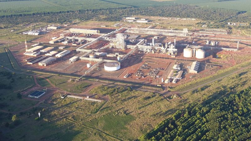 Vista aérea de complexo industrial de grande porte com solo avermelhado, tanques, estruturas metálicas e galpões, cercado por vegetação de cerrado. 
