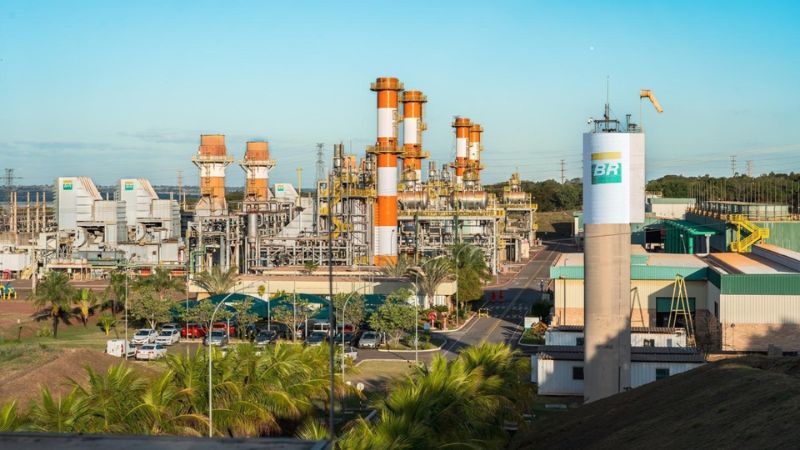 Vista externa da Usina Termelétrica de Três Lagoas, com chaminés listradas em laranja e branco, tanque com logo da Petrobras (BR) e vegetação ao redor, sob céu azul. 