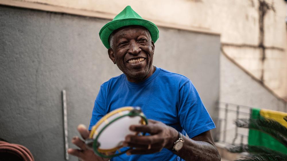 Homem tocando samba com um pandeiro. Ele veste as cores do Brasil e está com um sorriso no rosto.