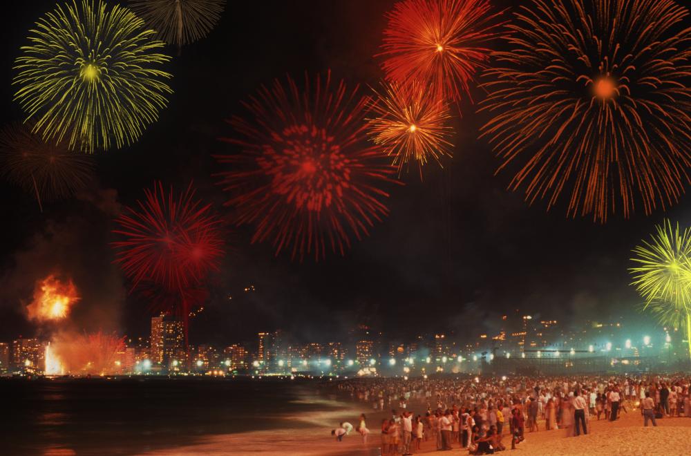 Pessoas reunidas na areia da praia observando fogos de artifício vermelhos e amarelos no céu noturno, com a orla de Copacabana iluminada ao fundo.