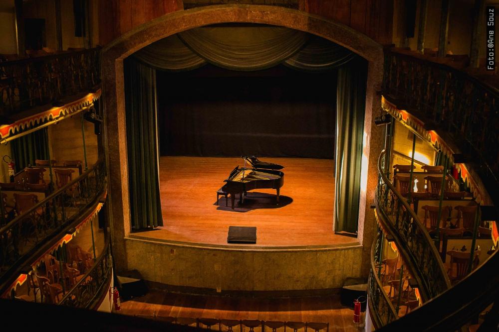 Vista aérea do interior de um teatro histórico, com um piano de cauda solitário e iluminado no centro do palco vazio. A moldura do proscênio é ornamentada e ladeada por cortinas verdes, e camarotes com balaustradas de ferro trabalhado ficam vazios dos dois lados.