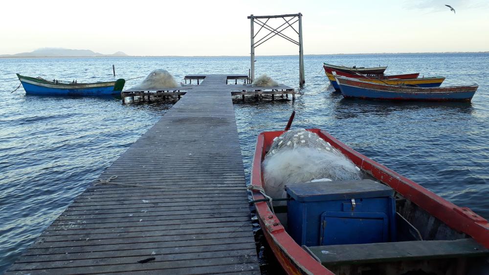 Pequenos barcos de pesca atracados em um trapiche de madeira sobre a água. No barco vermelho em primeiro plano, há redes e uma caixa azul de gelo.
