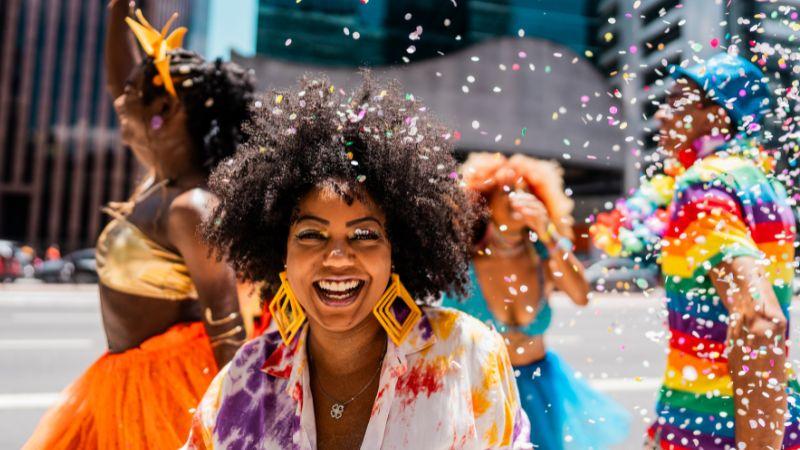 Foliões fantasiados celebram o Carnaval na rua sob uma chuva de confetes coloridos, destacando uma mulher sorrindo no centro.